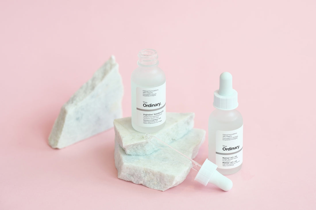 Skincare bottles rest on marble against a pink background.
